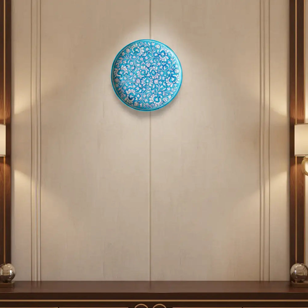 A round decorative wall plate with an intricate hand-painted white floral and vine pattern on a vibrant turquoise background.