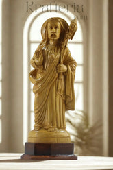 Statue of a religious figure with a staff on a wooden base, against a blurred architectural background.