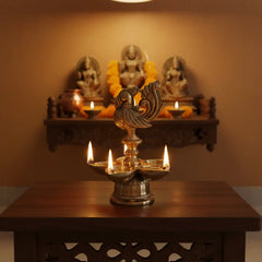 A golden brass five-wick oil lamp (Pancha Mukhi Diya) featuring a detailed peacock figure on top, with five small deepak bowls arranged around its base, against a light textured background.