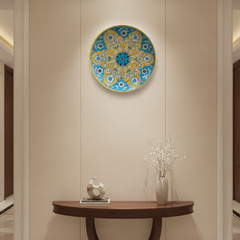 A round decorative wall plate with a vibrant yellow and teal floral medallion pattern, featuring white floral details and a central teal and maroon flower, framed by a yellow rim.