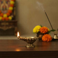 A small, golden brass diya with a decorative, intricately carved round base, holding a boat-shaped lamp on a white surface with a light-colored background.