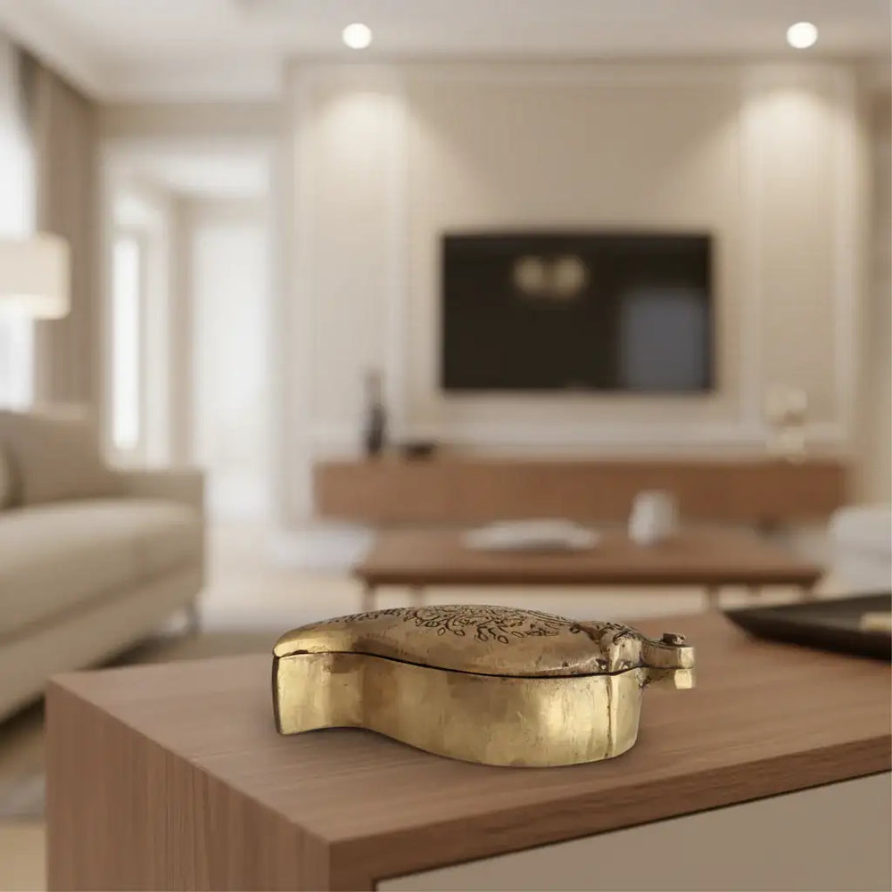 Top-down view of a small, ornate brass box in a mango or paisley shape, featuring detailed floral engravings and a small clasp/handle at one end, on a white surface.