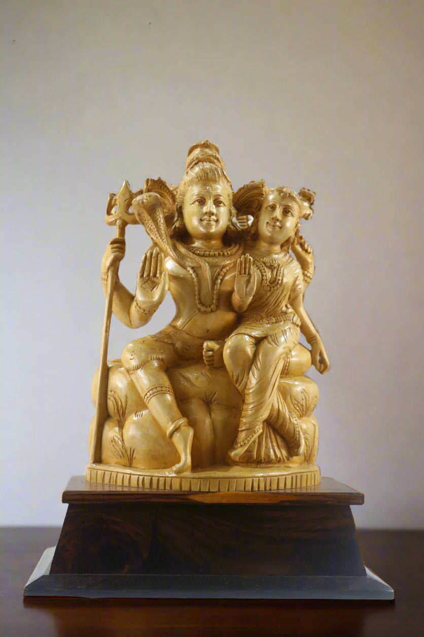 Gold-colored statue of a seated figure with another figure on a wooden base, displayed indoors.