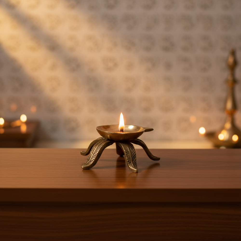 Brass diya stand with a shallow bowl and four decorative leaf-shaped feet on a white surface, with a light patterned background.