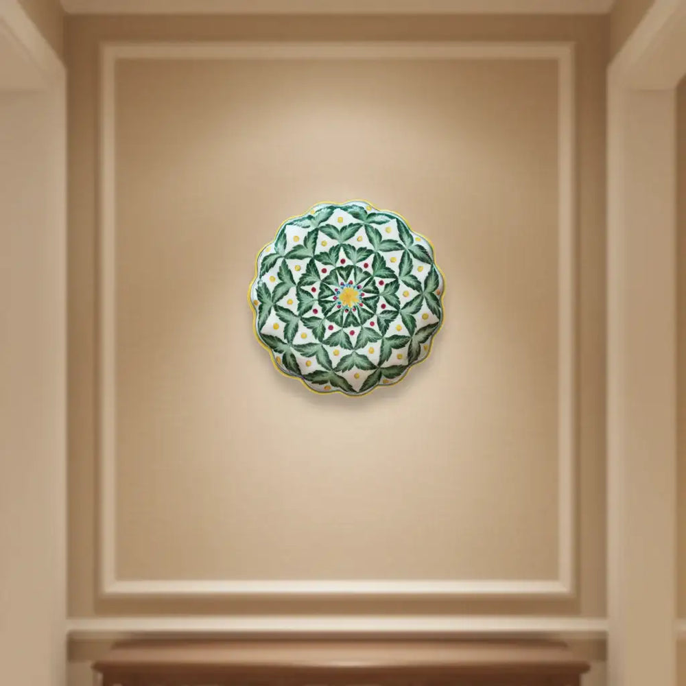 A decorative wall plate with a pattern of green leaves, small red dots, and yellow dots arranged around a yellow and blue starburst center, with a scalloped edge.