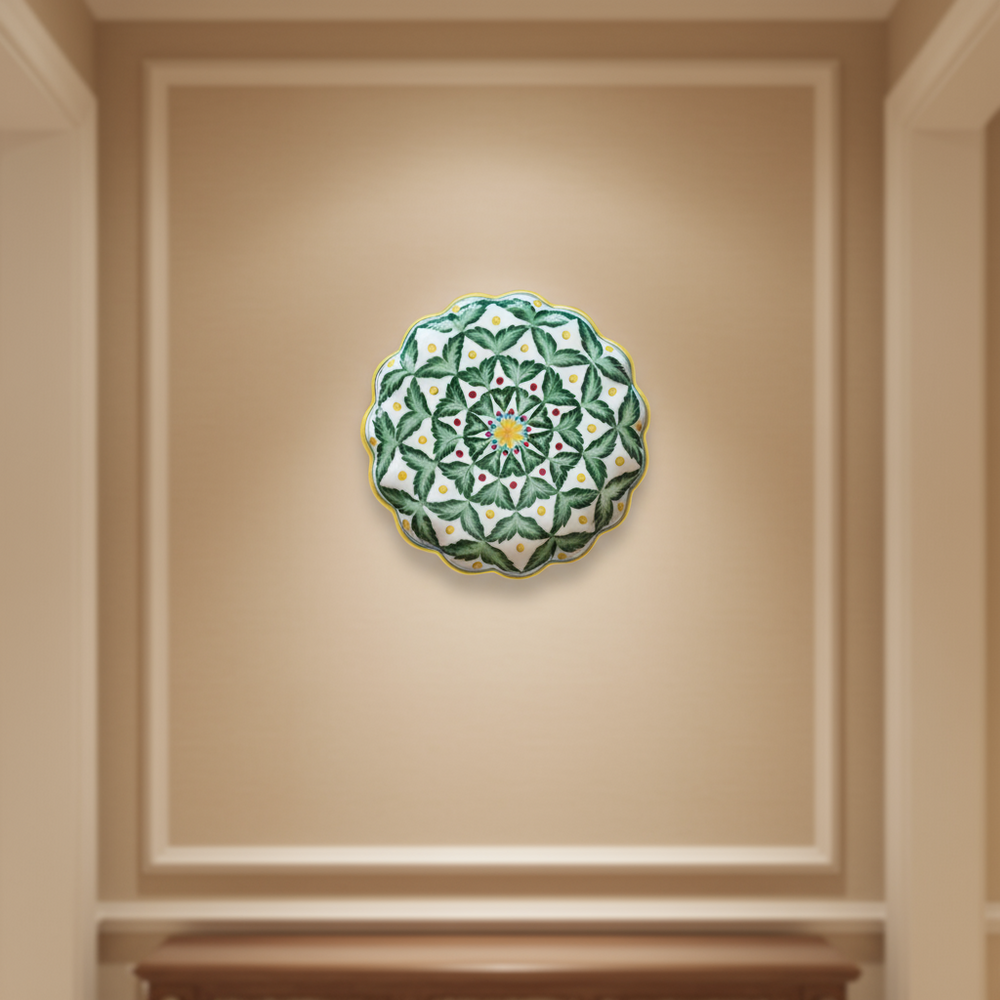 A decorative wall plate with a pattern of green leaves, small red dots, and yellow dots arranged around a yellow and blue starburst center, with a scalloped edge.