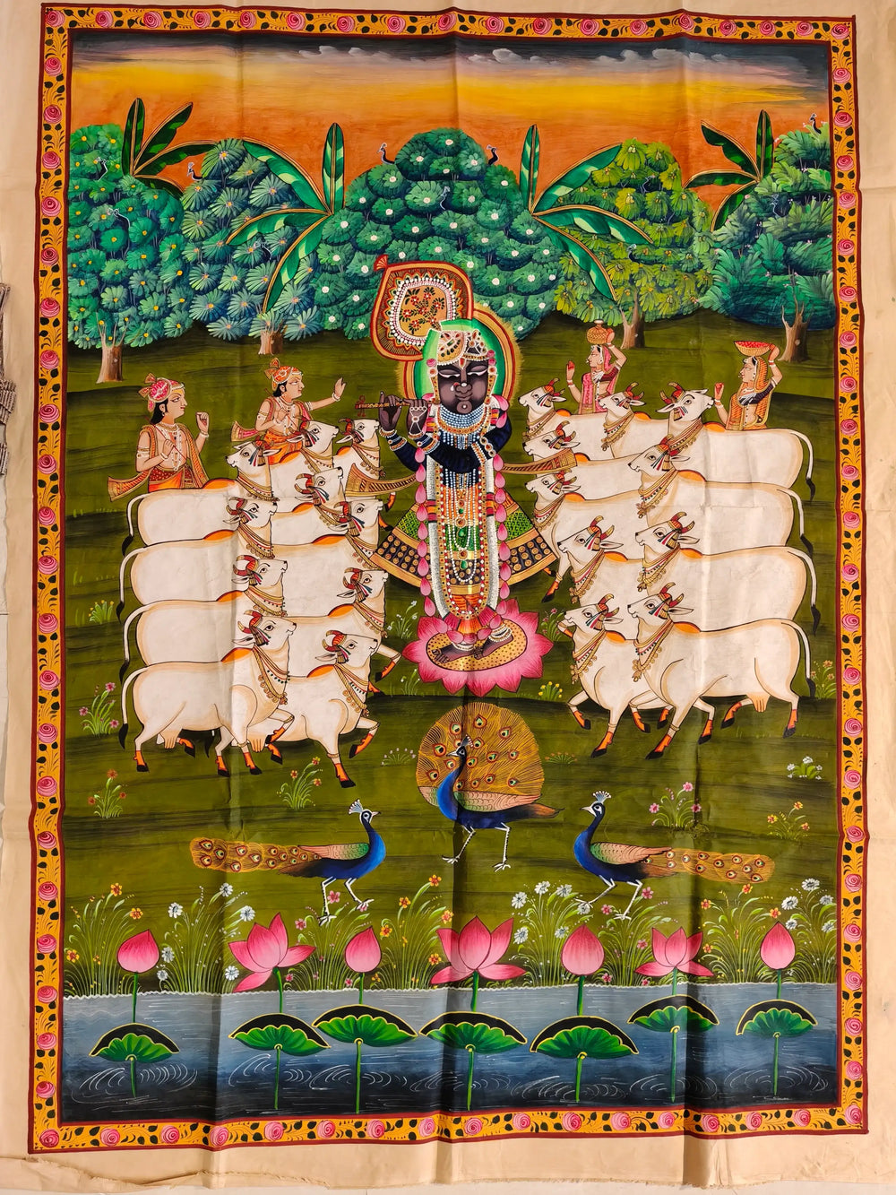 A traditional Indian Pichwai painting on a green background, showing Lord Krishna (Shrinathji) playing a flute, surrounded by gopis, a herd of white cows, peacocks, and a lotus pond at the bottom, all within a decorative border.