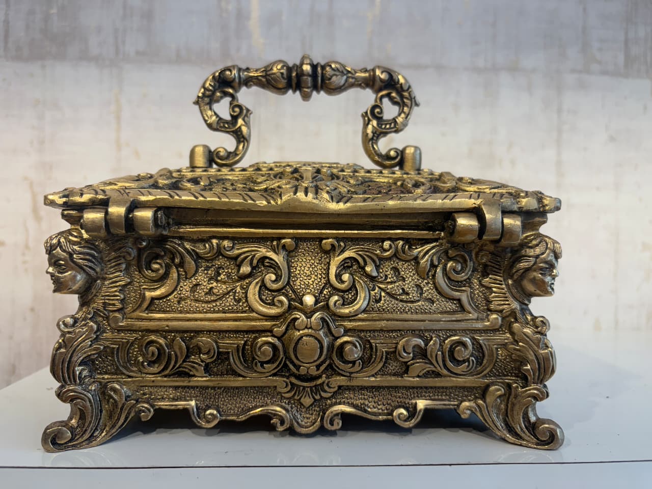 An ornate antique-style brass trinket box with a decorative handle, featuring intricate scrollwork and sculpted faces on its sides, sitting on a white surface.