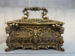 An ornate antique-style brass trinket box with a decorative handle, featuring intricate scrollwork and sculpted faces on its sides, sitting on a white surface.