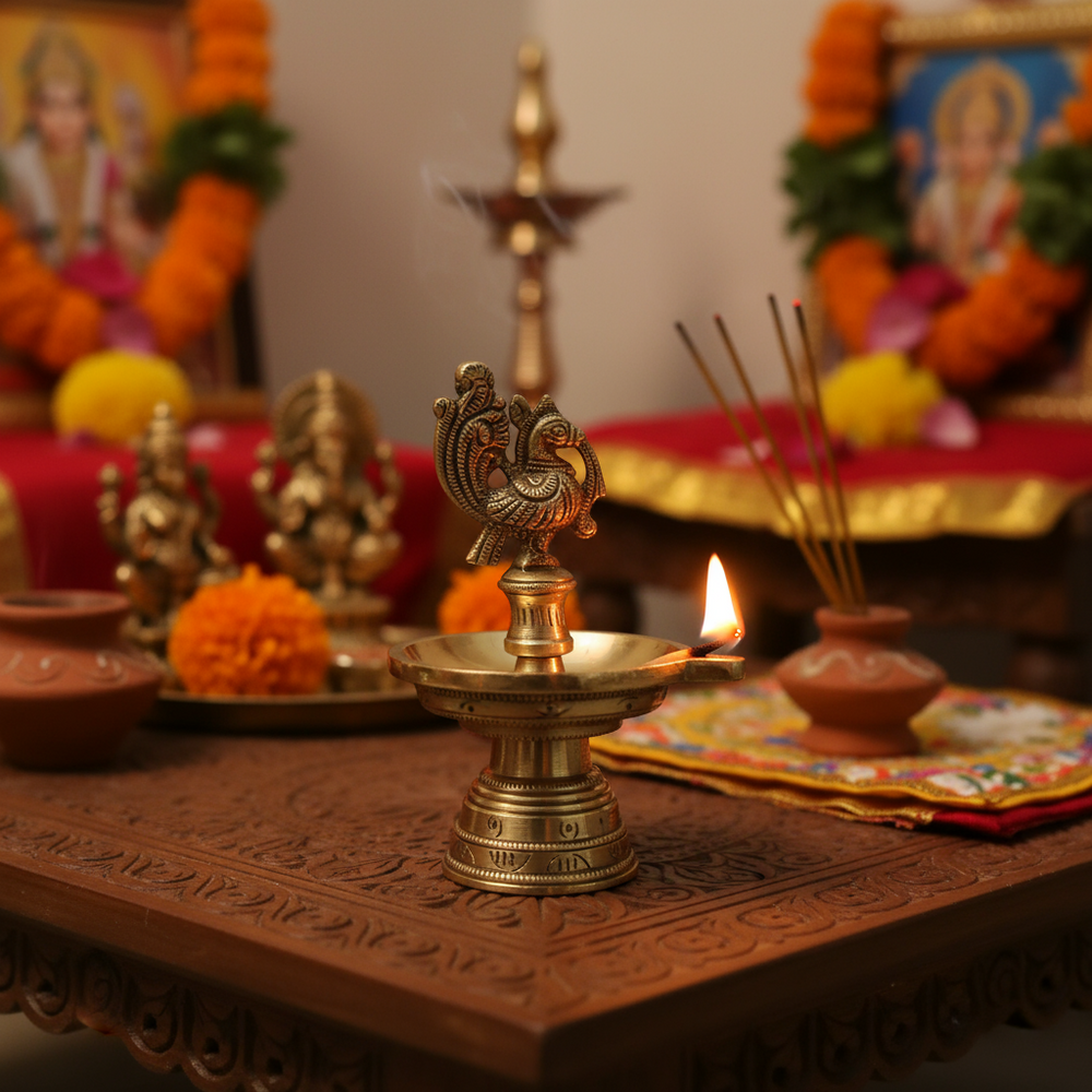 A golden brass traditional South Indian Kuthu Vilakku (oil lamp) with a wide base, a single deepak bowl, and an ornate peacock figure at the top, against a light textured background.
