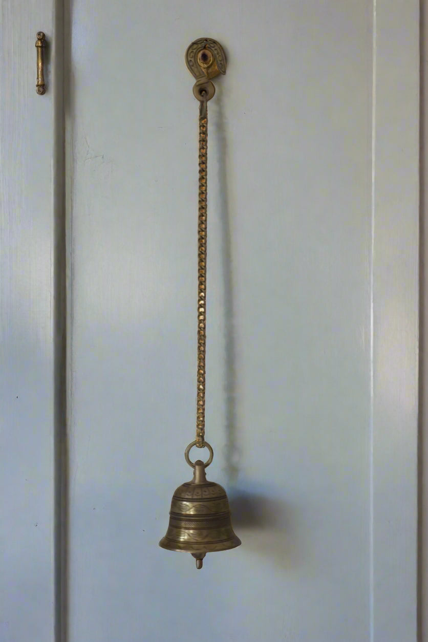 Decorative metal bell hanging in a sunlit room with intricate ceiling designs.