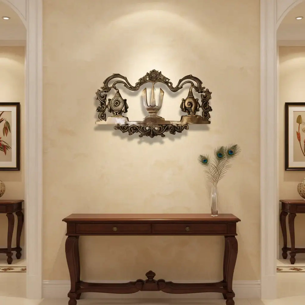 An ornate brass wall hanging displaying the U-shaped Venkateswara Namam (tilak) in the center, flanked by the Shankha (conch) and Chakra (discus) symbols, with decorative border work.