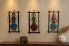 Three decorative wall hangings of women in colorful outfits on a beige wall.