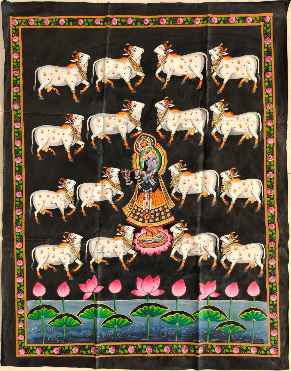 A traditional Indian Pichwai painting on a black background, depicting Lord Krishna (Shrinathji) playing a flute, surrounded by white cows, and a pond with pink lotus flowers at the bottom. The painting has a decorative border.