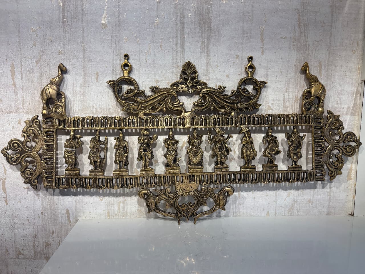 An ornate brass horizontal wall hanging featuring a row of ten standing Hindu deities or figures, flanked by decorative motifs and elephants, on a light background.