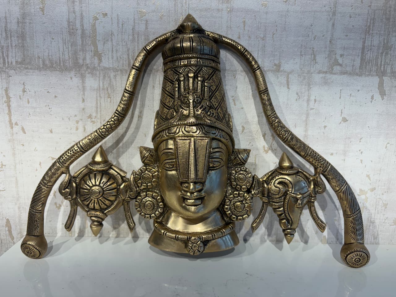 A detailed brass wall hanging depicting the face of Lord Venkateswara, adorned with a tall crown, traditional markings, and flanked by symbolic conch and discus motifs.