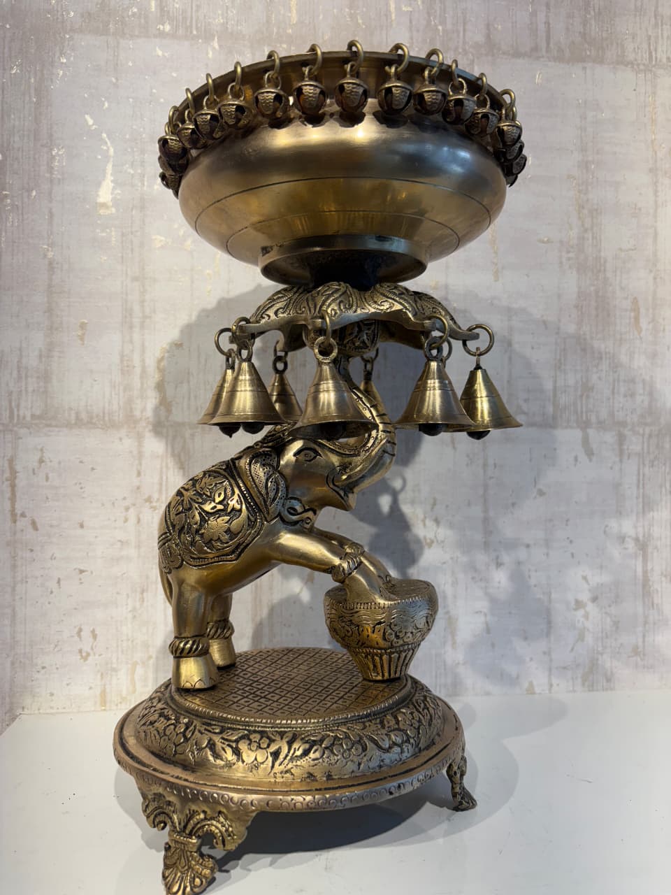 A detailed brass Urli (decorative floating bowl) with a prominent elephant base and numerous small brass bells hanging from the rim, set against a light background.