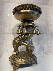 A detailed brass Urli (decorative floating bowl) with a prominent elephant base and numerous small brass bells hanging from the rim, set against a light background.