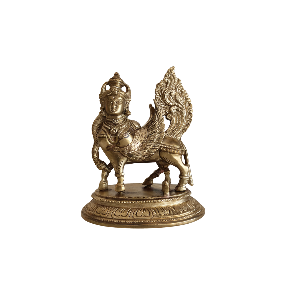 Front view of a shiny brass Kamadhenu Kalpavriksha cow statue with a human head, wings, and an ornate tail on a decorative